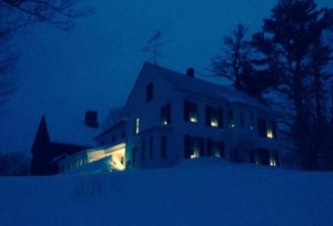 white colonial home atop snow covered hill with lights glowing in each window