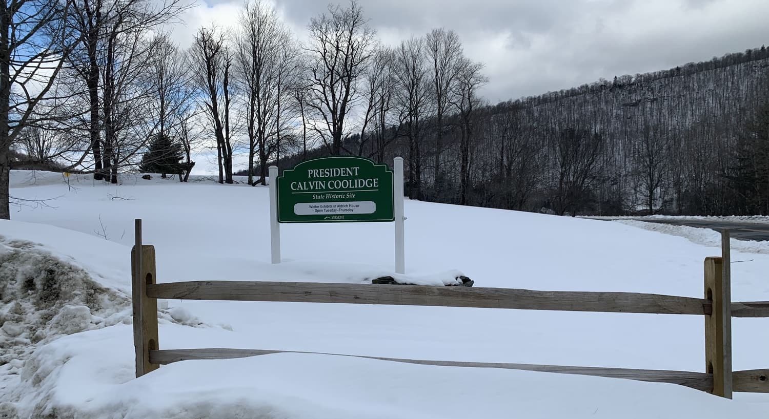 Green and white sign for President Calvin Coolidge State Historic Site surrounded by snow-covered ground and bare trees