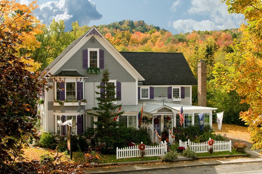 Front view of house painted gray and white with dark purple shutters surrounded by many trees with fall foliage