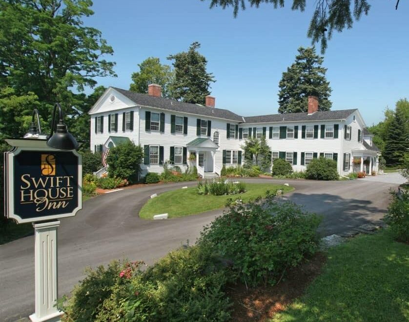 Front view of large house painted white with dark shutters surrounded by trees, shrubs and flowers