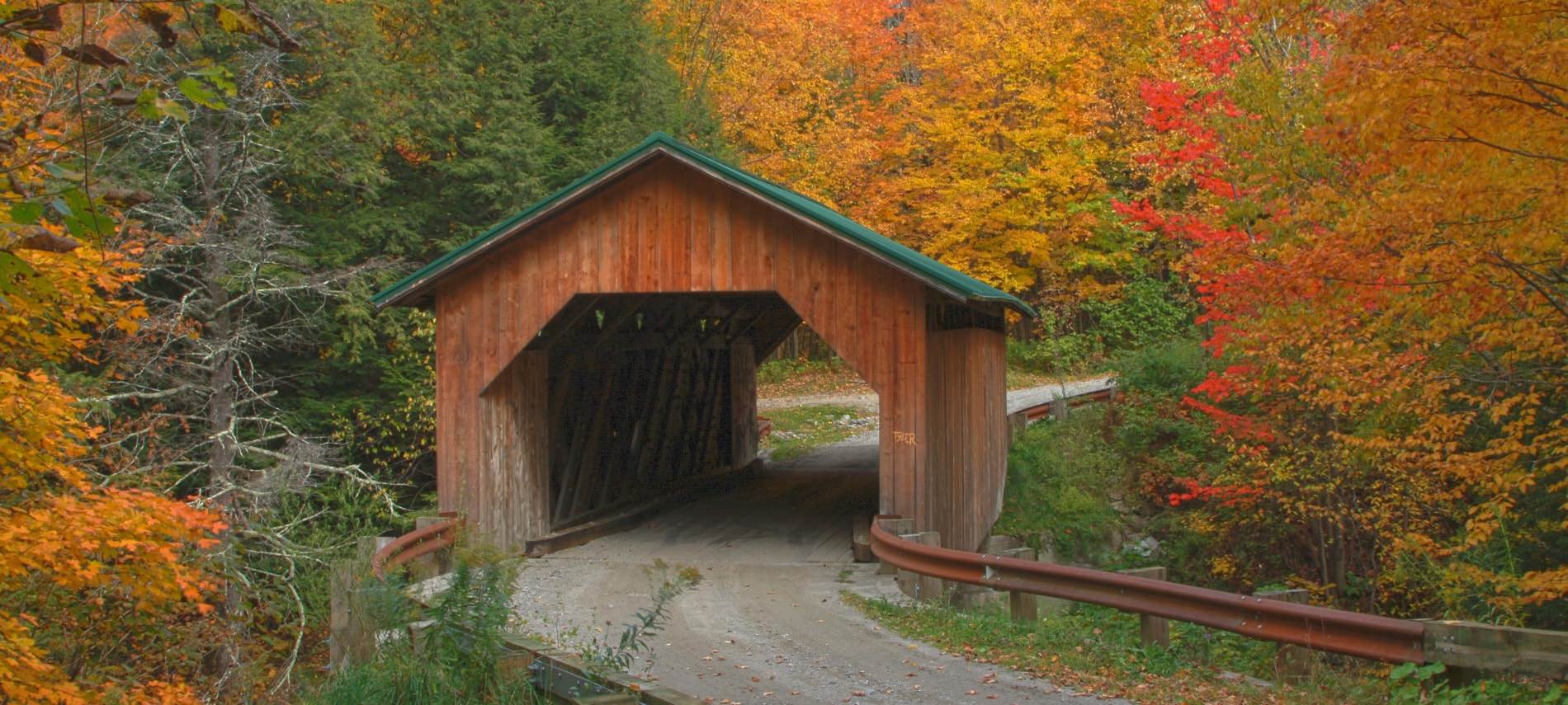 Cute wood covered bridge with green roof surrounded by trees filled with vibrant fall foliage
