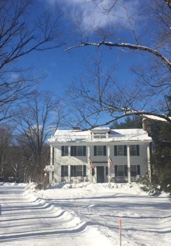 Exterior view of snowy lane leading to two-story white-sided inn amidst bare snow-covered trees and blue skies
